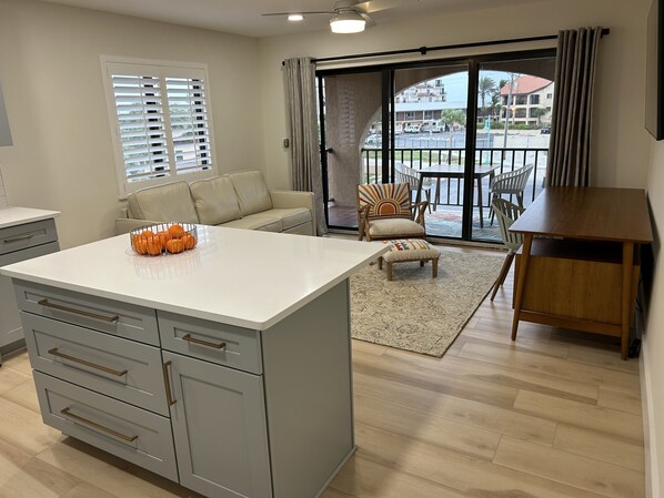 A modern kitchen with a central island, a dining area with a wooden table, and a view of the street through large windows. - Sunshine Swimmers Casita, 206 (Englewood)