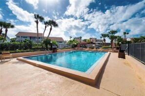 En rektangulær swimmingpool med fliseterrasse, omgivet af palmer og et hegn.