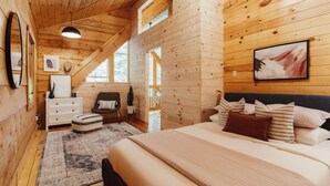 A wooden cabin bedroom with a bed, dresser, chair, and a large window. - Boutique All Wood Cottage in the Woods (Lanark)