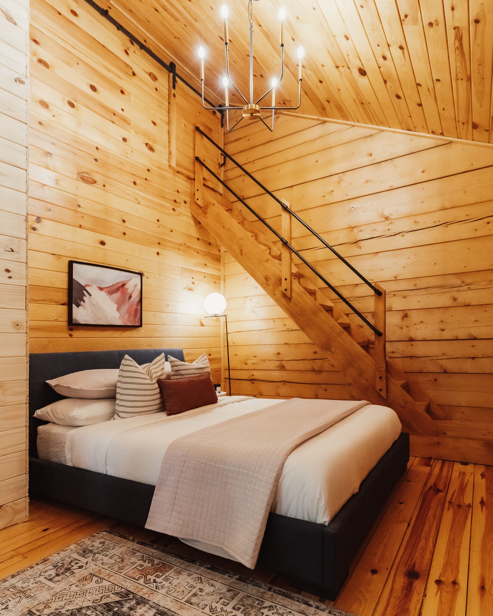 Ein Schlafzimmer einer Holzhütte mit Bett, Kissen, einer Nachttischlampe und einer Treppe.