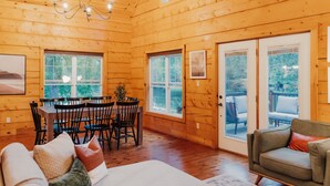 A cozy wooden cabin interior with a dining area, a sofa, and a sliding glass door leading to an outdoor deck. - Boutique All Wood Cottage in the Woods (Lanark)