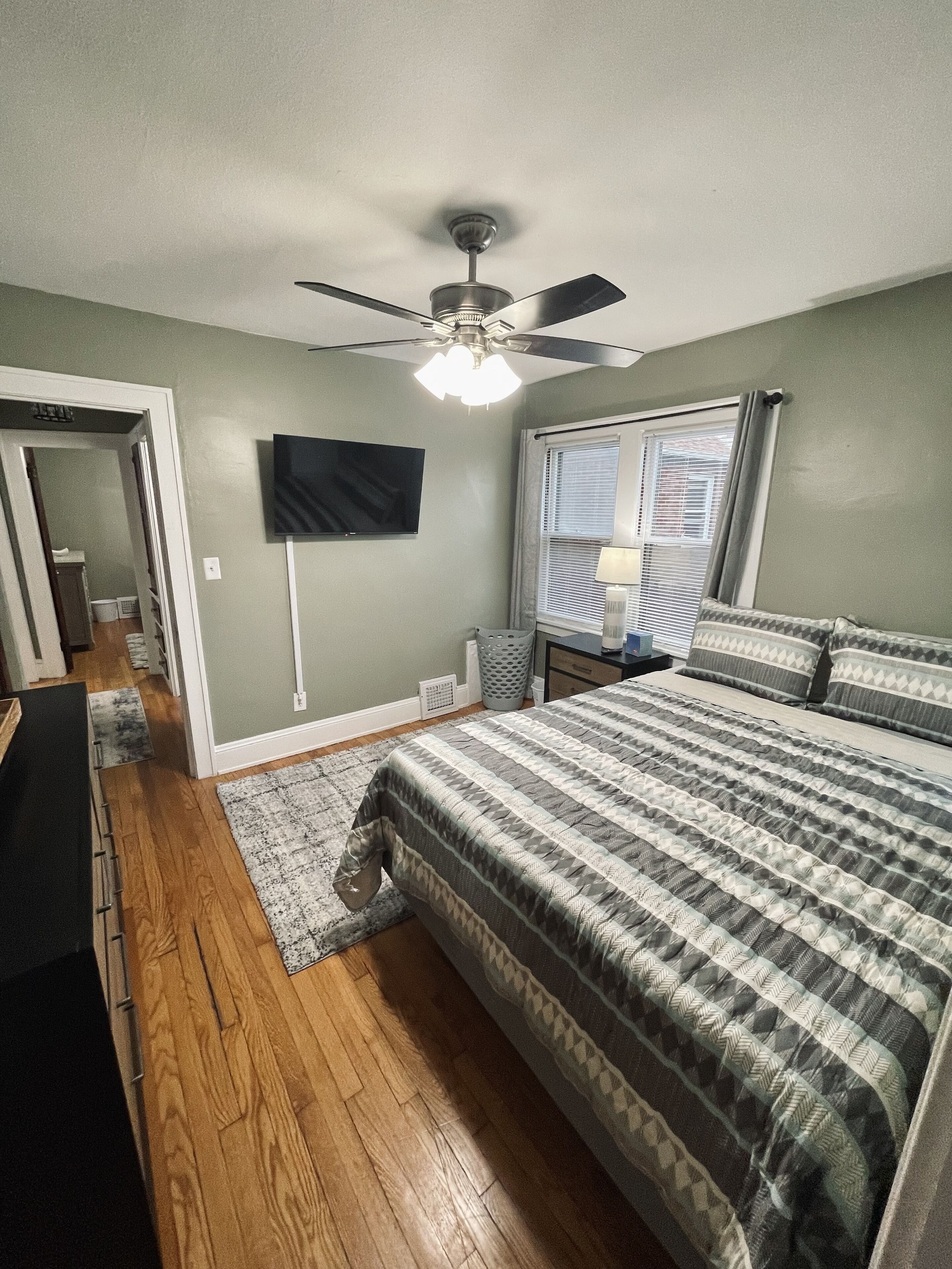 A bedroom with a bed, a ceiling fan, a TV mounted on the wall, a window with blinds, and a door leading to another room.