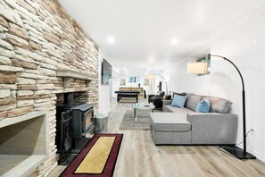 Una sala de estar moderna con pared de piedra, chimenea, sofá gris y una zona de comedor al fondo.