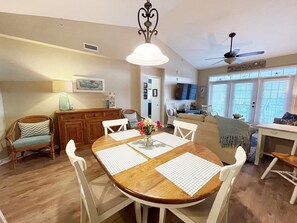 Un espace repas comprenant une table en bois, des chaises blanches, un lustre et un ventilateur de plafond.