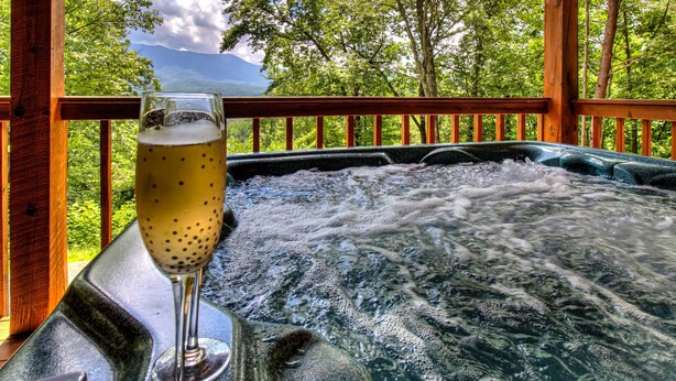 A hot tub with a glass of beer on a wooden deck with a view of trees and mountains. - Romantic and Cozy! 2 Bed 2 Bath True Log Cabin close to Park and Attractions!  (Gatlinburg)
