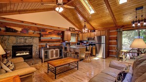 A cozy cabin interior with a stone fireplace, wooden furniture, a kitchen area, and a large window offering a view of the outdoors. - Romantic and Cozy! 2 Bed 2 Bath True Log Cabin close to Park and Attractions!  (Gatlinburg)