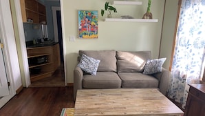Un salon avec une table basse en bois, un canapé agrémenté de coussins à motifs, un tableau coloré accroché au mur et, au fond, un coin cuisine.