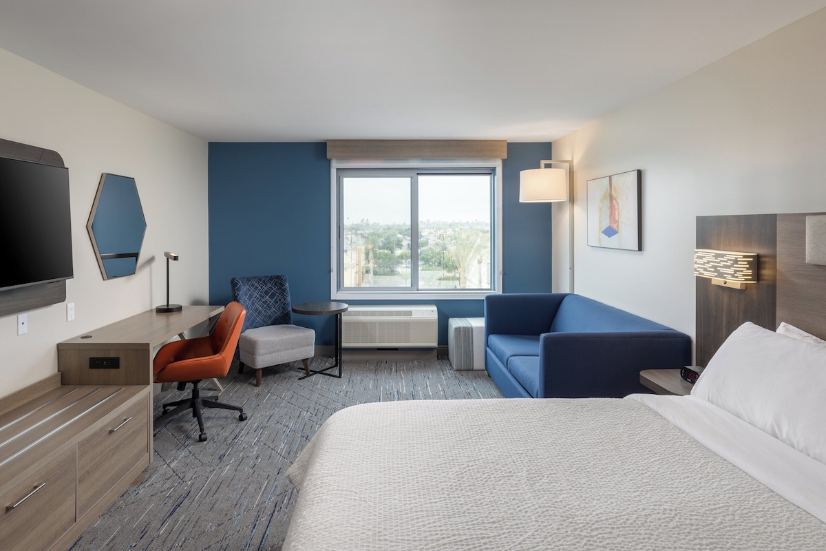 A hotel room with a bed, a desk, a chair, a sofa, a TV, and a window with a view.