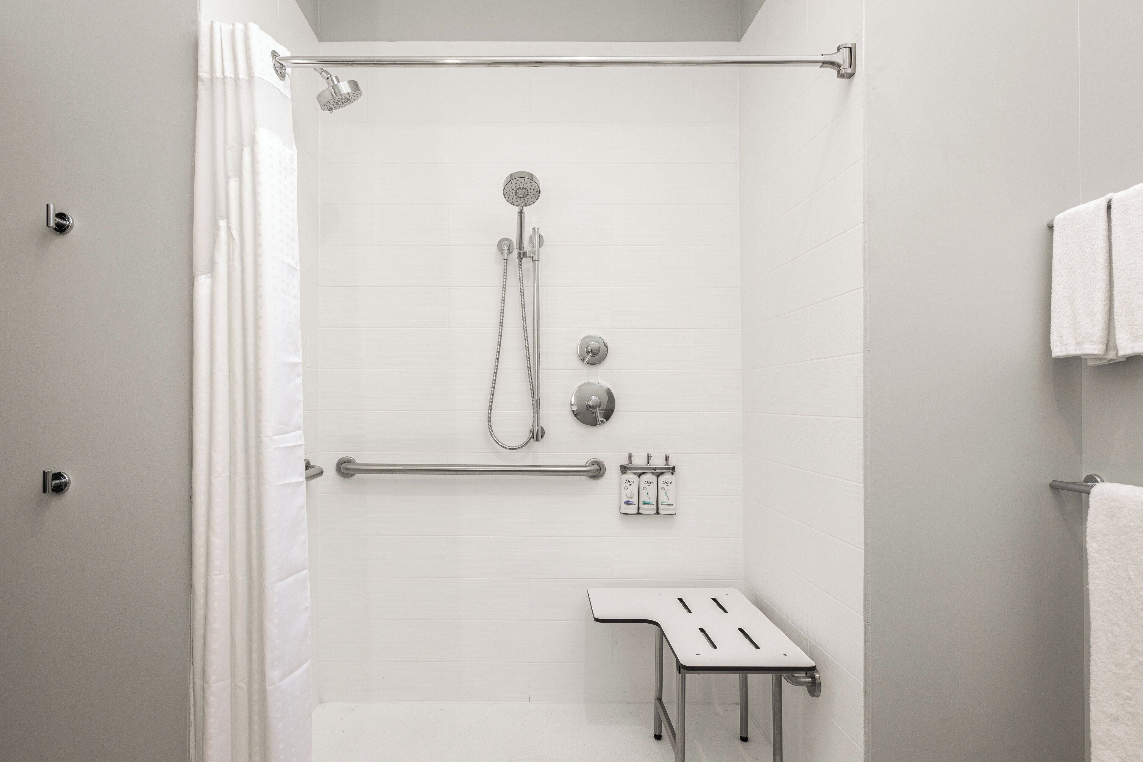 A modern shower area with a handheld showerhead, a handheld shower attachment, and a wall-mounted faucet. There is a grab bar for support, a small table with a towel, and a towel rack with a towel hanging on it.