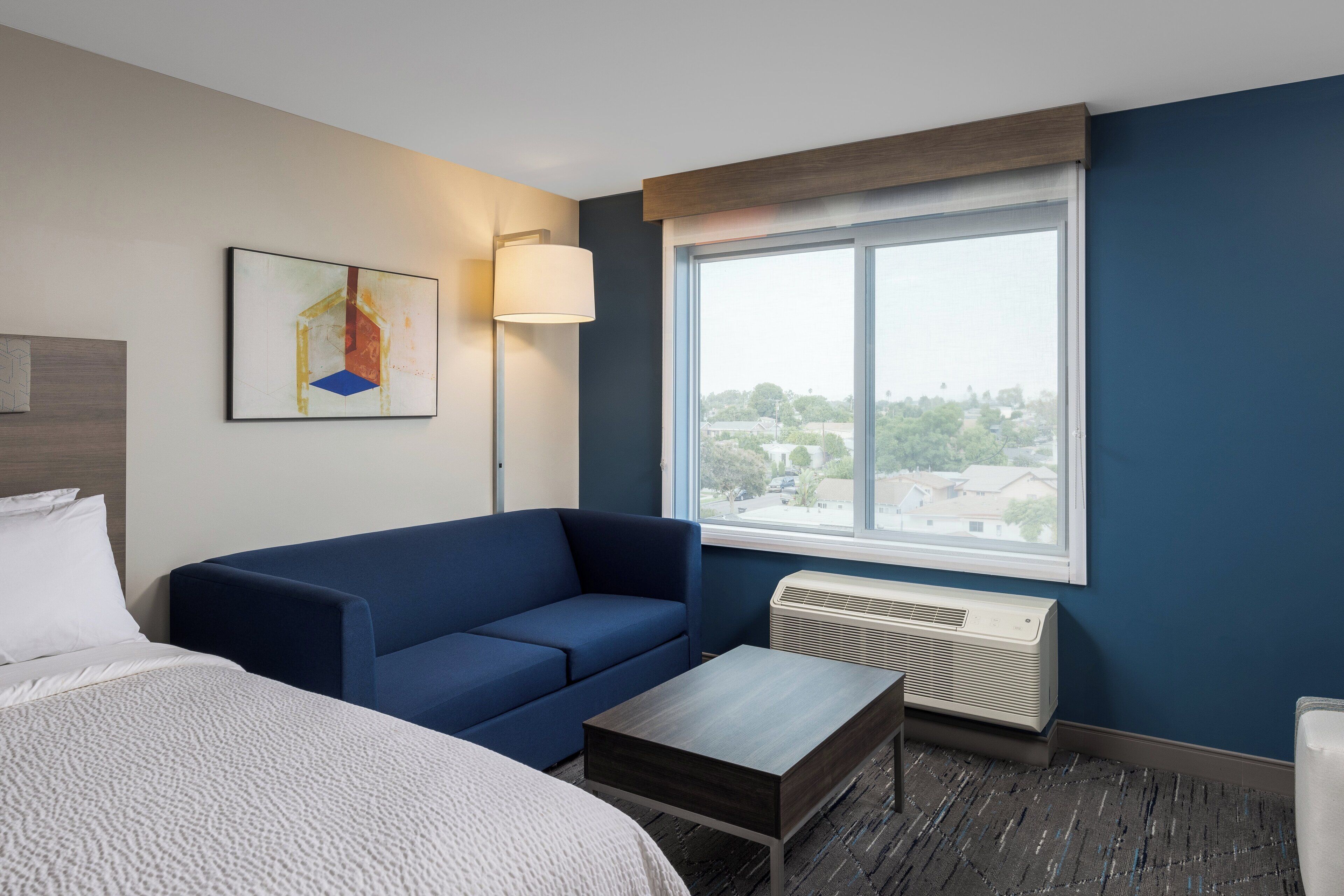 A hotel room with a bed, a blue sofa, a small table, a window with a view, and a wall-mounted air conditioner.