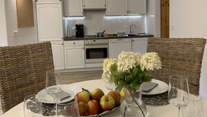 On aperçoit un coin repas avec des chaises en osier, une table dressée avec des assiettes, des verres et un saladier rempli de fruits. Au fond, on distingue une cuisine avec des meubles blancs et un fourneau.