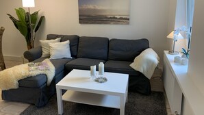 Un salon avec un canapé gris foncé, des coussins blancs, une table basse blanche et un tableau représentant une scène de plage accroché au mur.