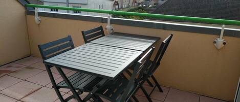 A balcony with a table and chairs, a green railing, and a view of buildings in the background.