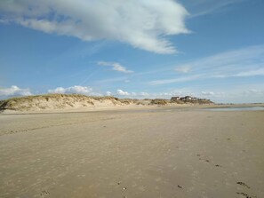 Uma extensa praia de areia com pegadas, dunas e um edifício ao longe.