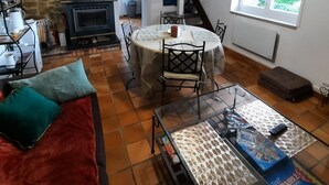 A cozy living room with a fireplace, a dining area with a table and chairs, a sofa with cushions, and a window with a view of greenery.