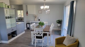 A modern kitchen with white cabinets, a dining area with a wooden table and white chairs, and a yellow armchair. - Bright, Modern Park Forest Home, 2.5m to Penn State! Breakfast included.  (State College)