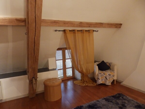 A room with a wooden beam ceiling, a window with blinds, a chair with a patterned cover, and a small stool. - Renovated 150-year-old building, close to Chateau de Menou. (Menou)
