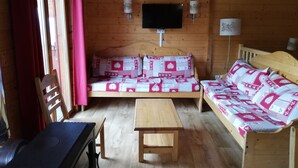 A cozy cabin-style room with a wooden interior, a sofa, a bed, a table, and a TV mounted on the wall. - Chalet 8 pers - 50m from the slopes (Valmeinier)