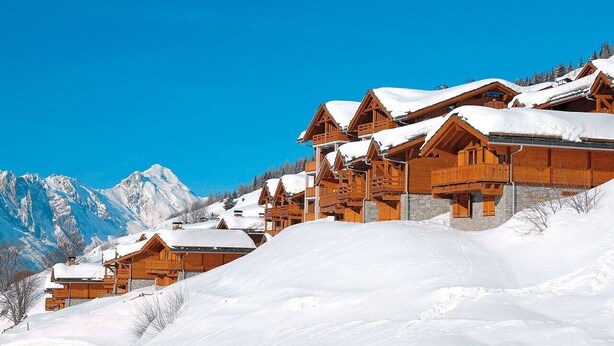 A row of wooden chalets with snow-covered roofs, set against a backdrop of snow-covered mountains and clear blue sky. - Chalet 8 pers - 50m from the slopes (Valmeinier)