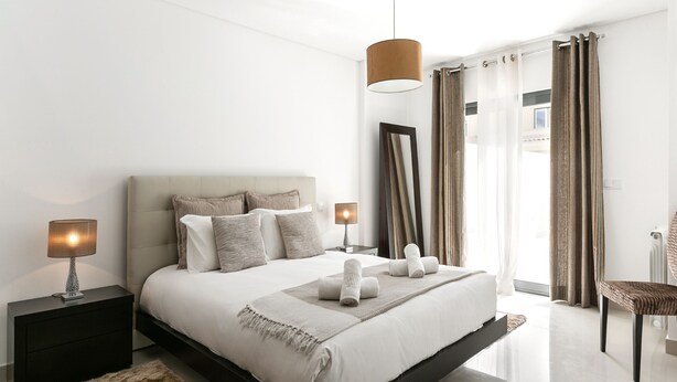 A modern bedroom with a large bed, bedside tables with lamps, a mirror, and a view of the outdoors. - Caminho da Praia - Pool & Beach Area (Peniche)