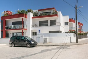Un immeuble moderne à plusieurs étages, doté de balcons, avec une voiture garée devant et un ciel bleu dégagé.
