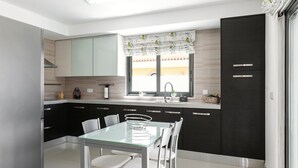 A modern kitchen with a glass dining table, white chairs, black cabinets, and a window with a curtain. - Caminho da Praia - Pool & Beach Area (Peniche)