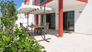A modern outdoor seating area with a table and chairs, a white umbrella, and a glass balustrade overlooking a building. - Caminho da Praia - Pool & Beach Area (Peniche)