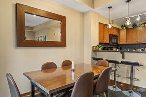 A dining area with a wooden table, four chairs, a large mirror, and a kitchen with wooden cabinets and bar stools.