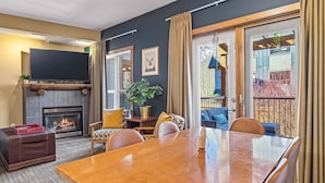 A spacious living room with a fireplace, a large flat-screen TV, and a dining area with a wooden table and chairs. - Family & Group Oasis: Hot Tub, AC, Fireplace, 2 Patios, Walk to Food & Shops (Canmore)