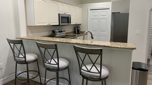 A kitchen with white cabinets, a granite countertop, a stainless steel refrigerator, and three bar stools with cushioned seats. - Coastal Tranquility in Quiet, Gated Community (Rotonda West)
