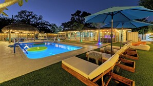 Zona de piscina con sillas de descanso y sombrillas, rodeada por una valla y árboles.