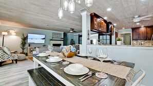 Una cocina moderna con armarios de madera, un comedor con una mesa larga dispuesta para cuatro personas y una sala de estar con sofá y televisión.