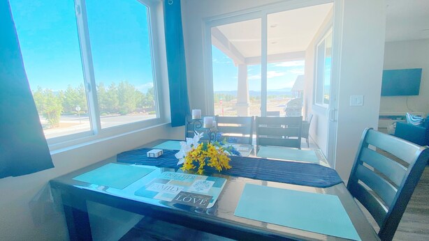 A room with a dining table set for two, a vase with yellow flowers, and a view of the outdoors through large windows. - Modern 3 BD House in Cottonwood Downtown, 20 mins to Sedona.  (Cottonwood)