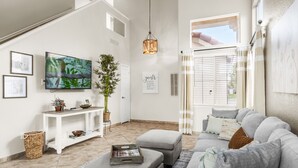 Una sala de estar con un sofá gris, una mesa de centro, un televisor, una lámpara colgante y un ventanal con cortinas.