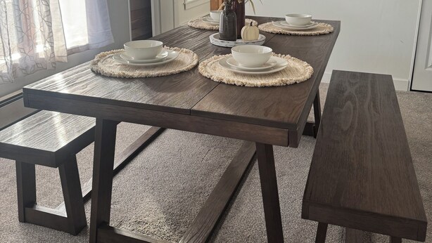 Un comedor con una mesa de madera dispuesta para cuatro personas, rodeado de un suelo alfombrado y una escalera.