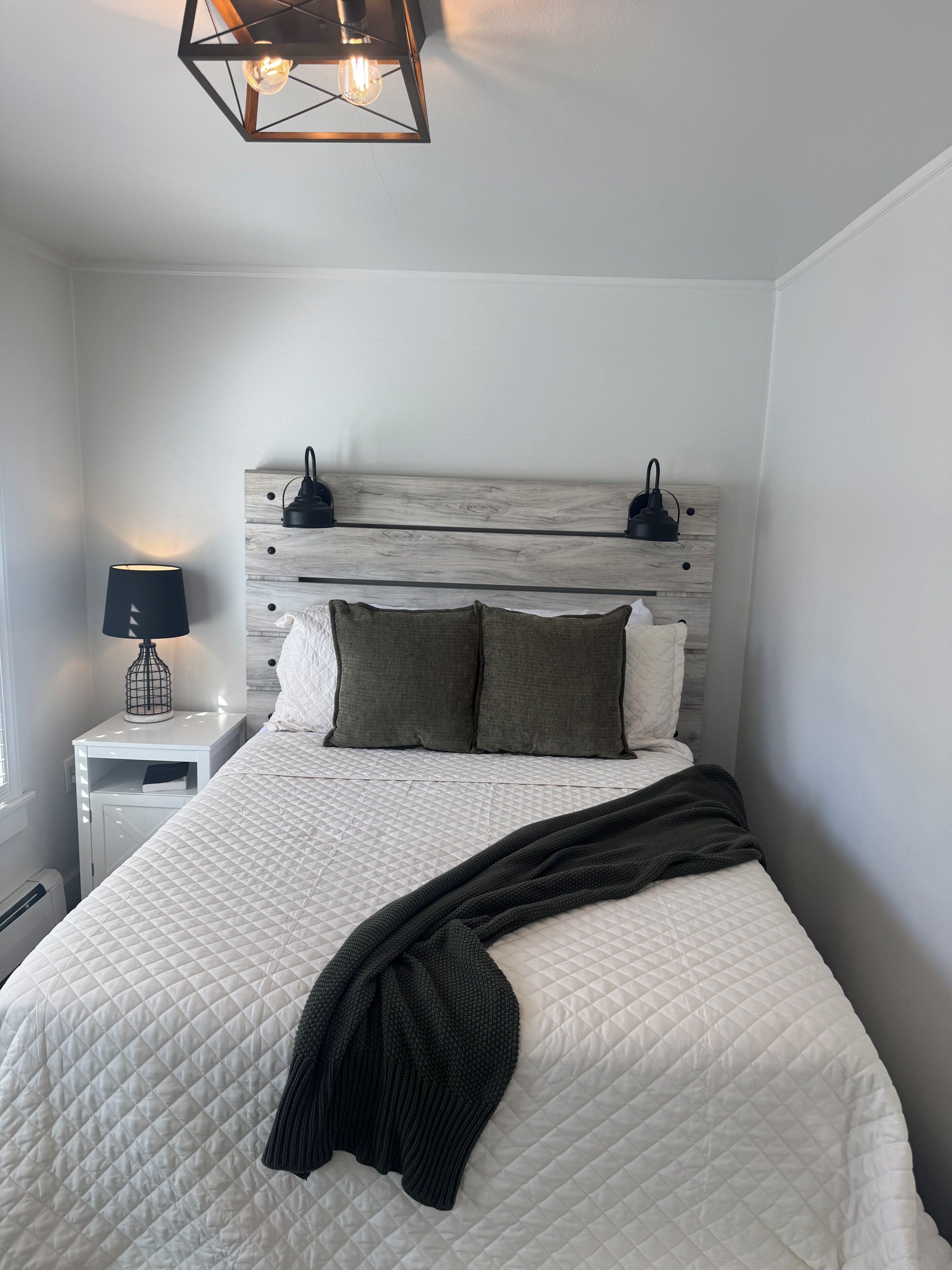 A neatly made bed with a quilted white duvet, a dark throw blanket, and two pillows. There is a bedside lamp on a white nightstand beside the bed.
