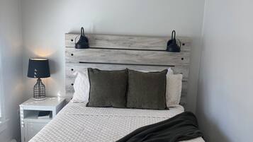 A neatly made bed with a quilted white duvet, a dark throw blanket, and two pillows. There is a bedside lamp on a white nightstand beside the bed.