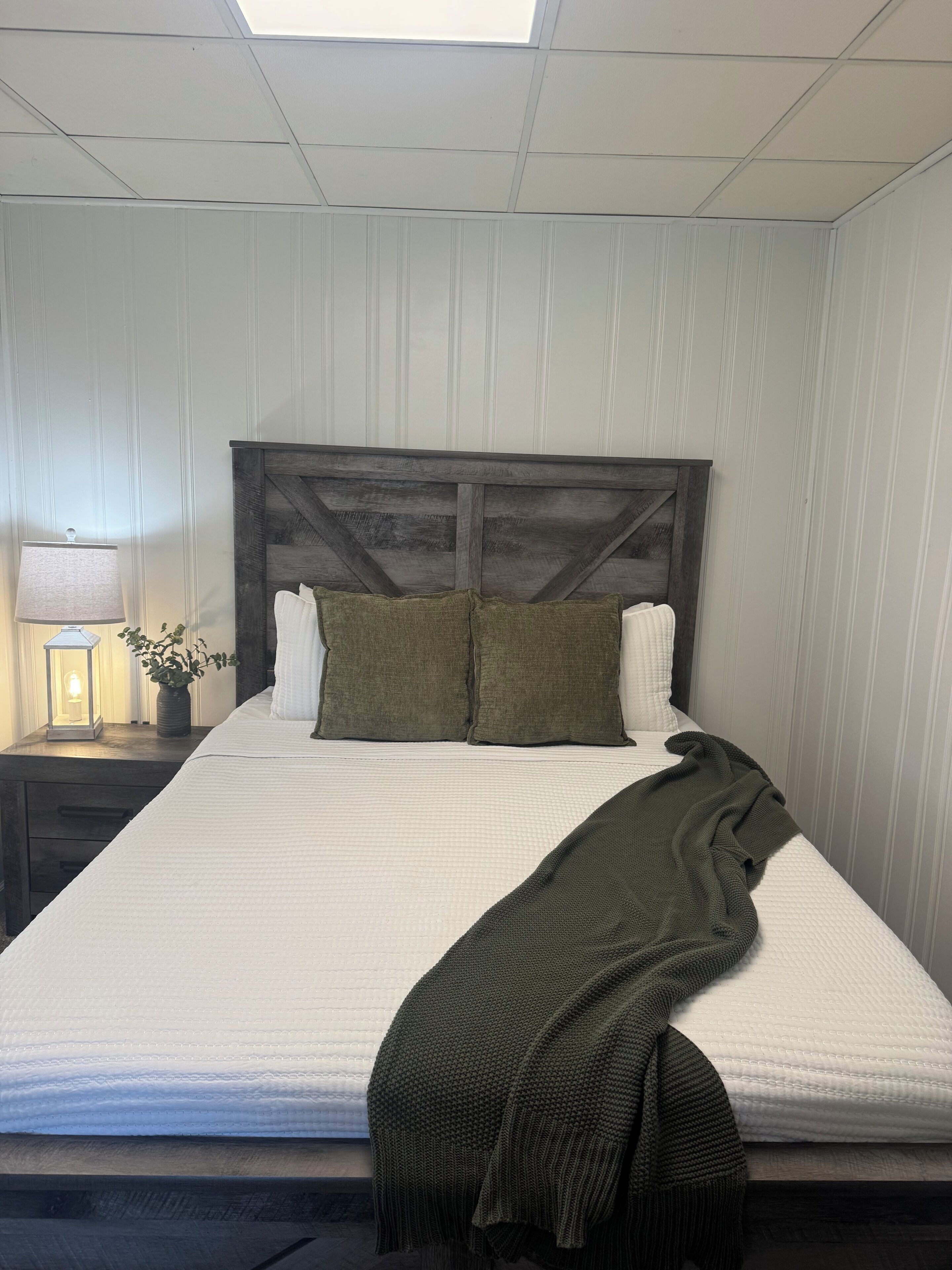 A neatly made bed with a wooden headboard, a bedside table with a lamp and a plant, and a window with a view of the sky.