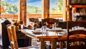 Une table de salle à manger en bois, mise avec des assiettes, des verres et un vase de fleurs. La table est placée près de grandes fenêtres qui offrent une vue sur un paysage montagneux.