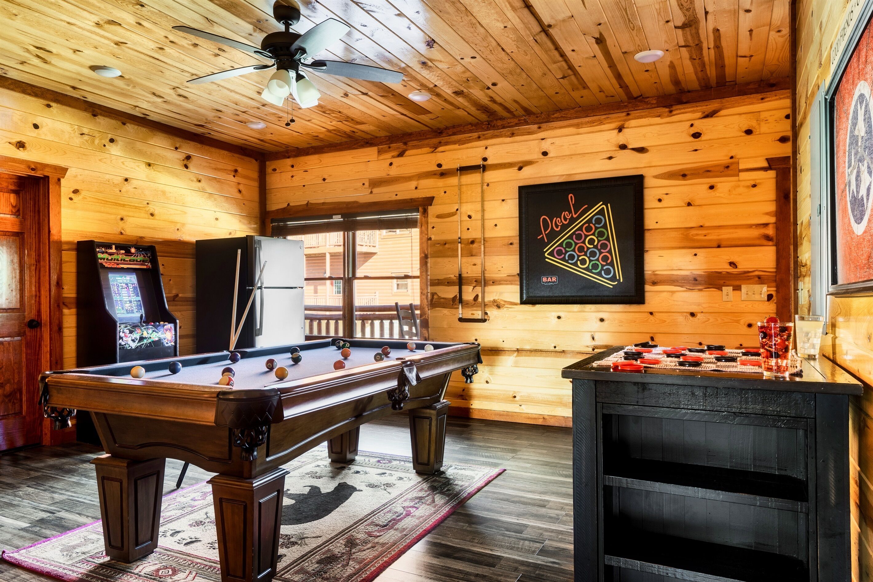 A pool table, foosball table, and arcade games in a wooden room with a ceiling fan.