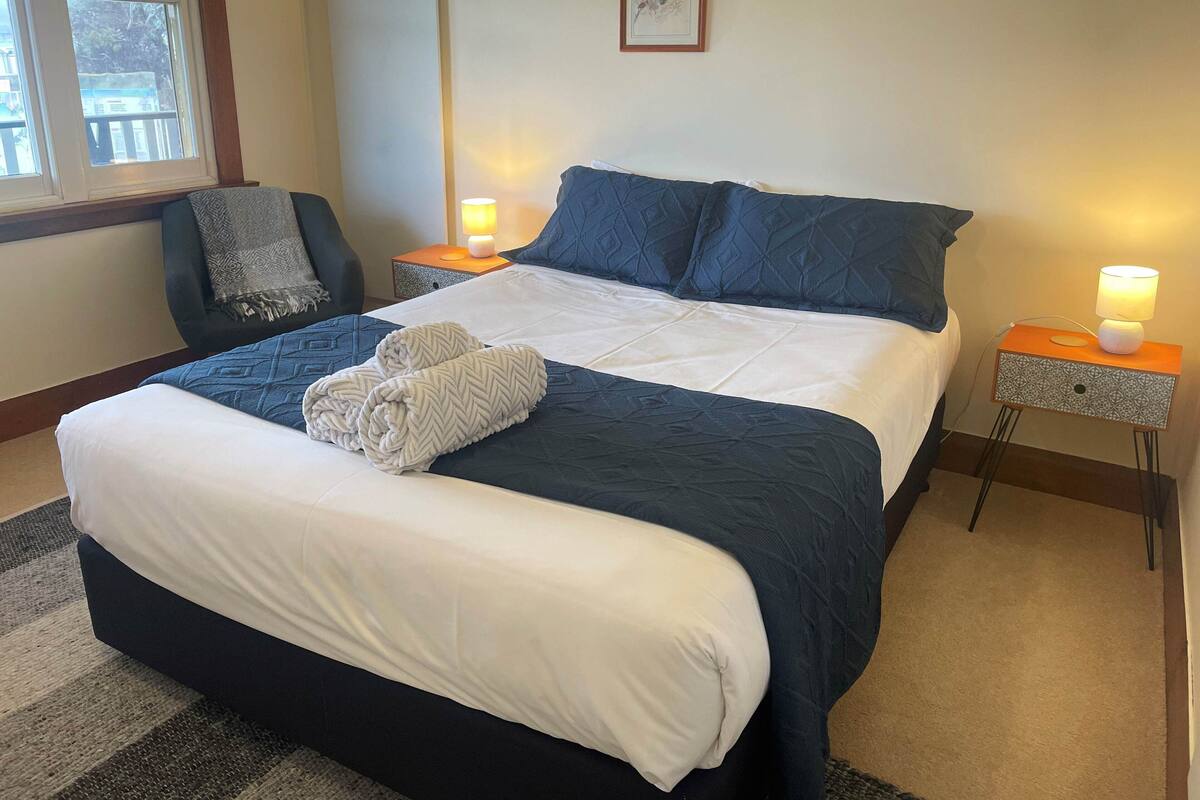 A neatly made bed with a white comforter and two folded towels, a dark blue quilted headboard, a chair with a patterned throw, and two bedside tables with lamps.