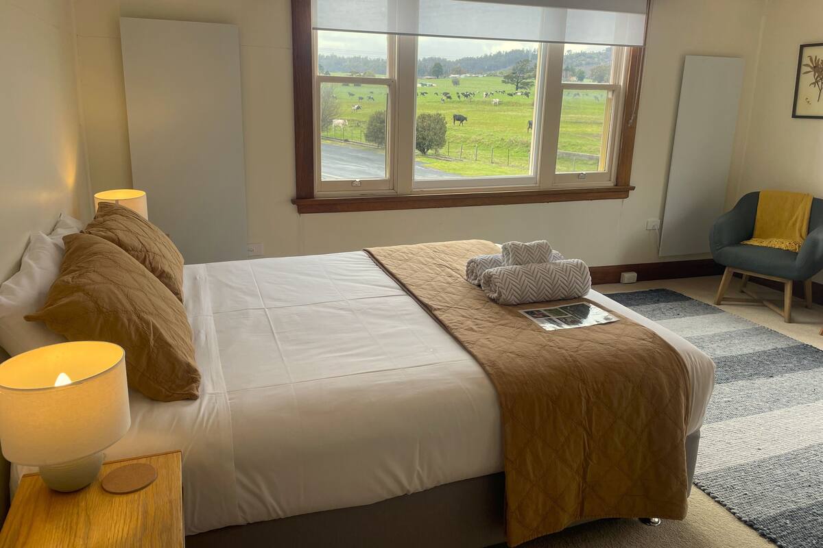 A bedroom with a large bed, a window with a view of a green field, a chair, and a lamp.
