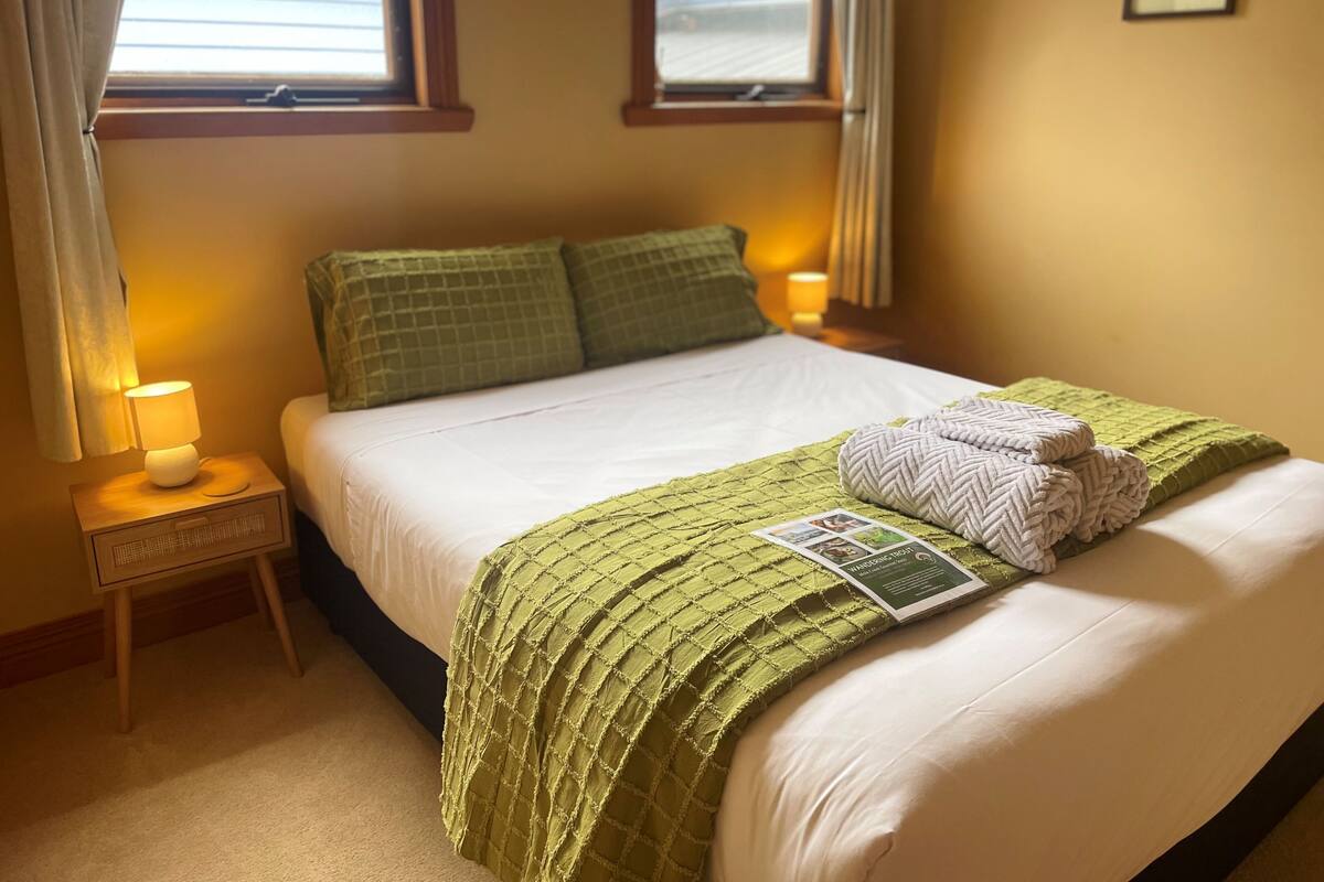 A neatly made bed with a green blanket, pillows, and a folded towel. There is a bedside lamp on a wooden nightstand.