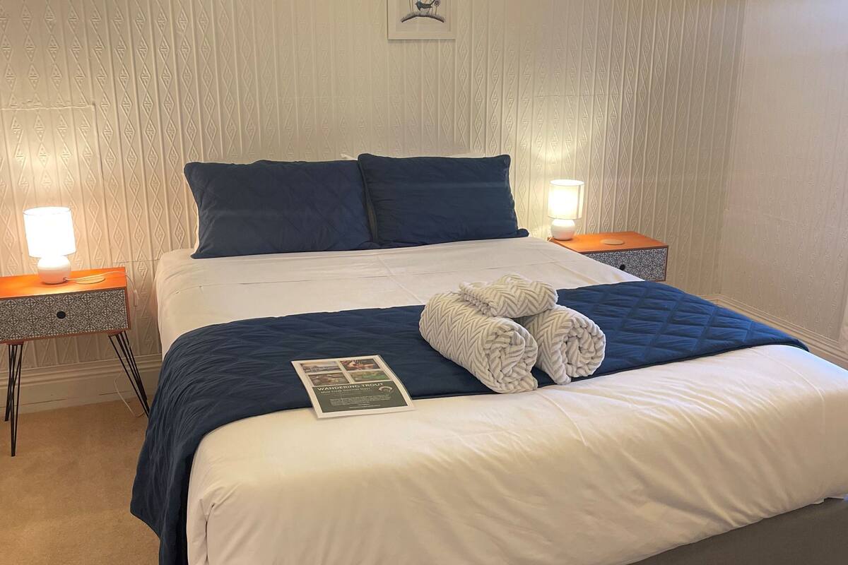 A neatly made bed with a blue and white duvet, two pillows, and a folded towel on the bed. There is a framed picture on the wall above the bed.