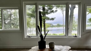 Una habitación con un gran ventanal con vista a un lago, una mesa de madera rústica con un libro y una taza, y un florero con plantas altas.
