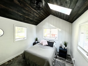 Ein Schlafzimmer mit einem Bett, zwei Nachttischen, einem Wandventilator und zwei Fenstern mit Jalousien.