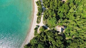 An aerial view of a coastal property with a beach, dense greenery, and a house surrounded by trees.