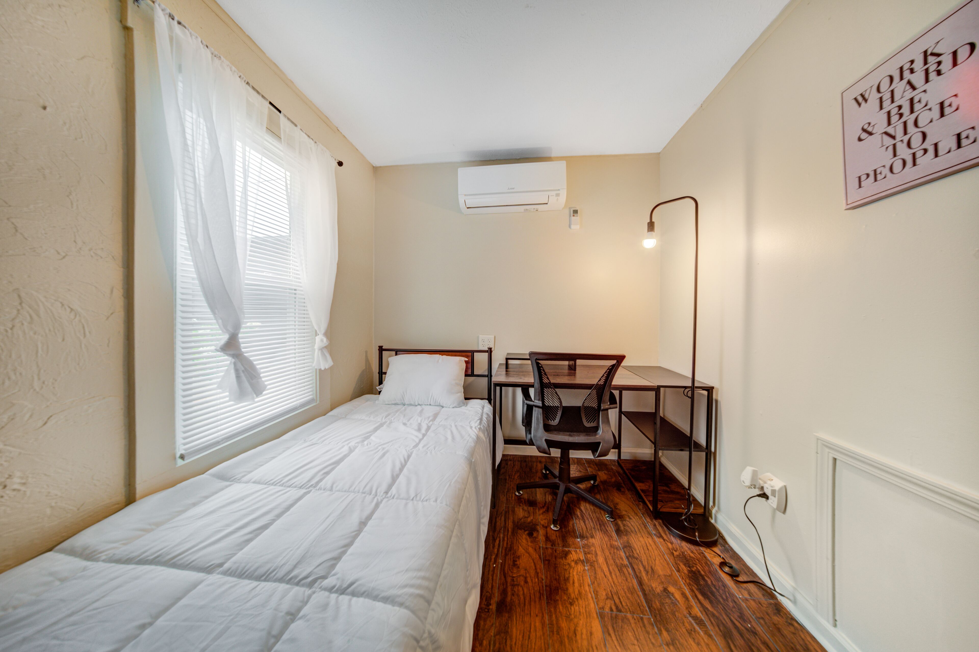 A small, well-lit bedroom with a single bed, a desk with a chair, and a wall-mounted air conditioner.