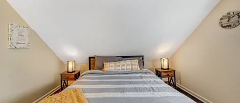 A bedroom with a sloped ceiling, a bed with striped bedding, two bedside tables with lamps, and a wall clock.
