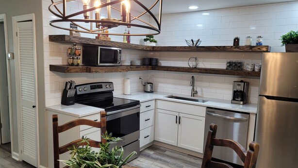 A modern kitchen with white cabinets, a stainless steel refrigerator, a built-in oven, and a microwave. There is a wooden dining table with chairs and a potted plant. - Chic, newly renovated townhome (North Charleston)